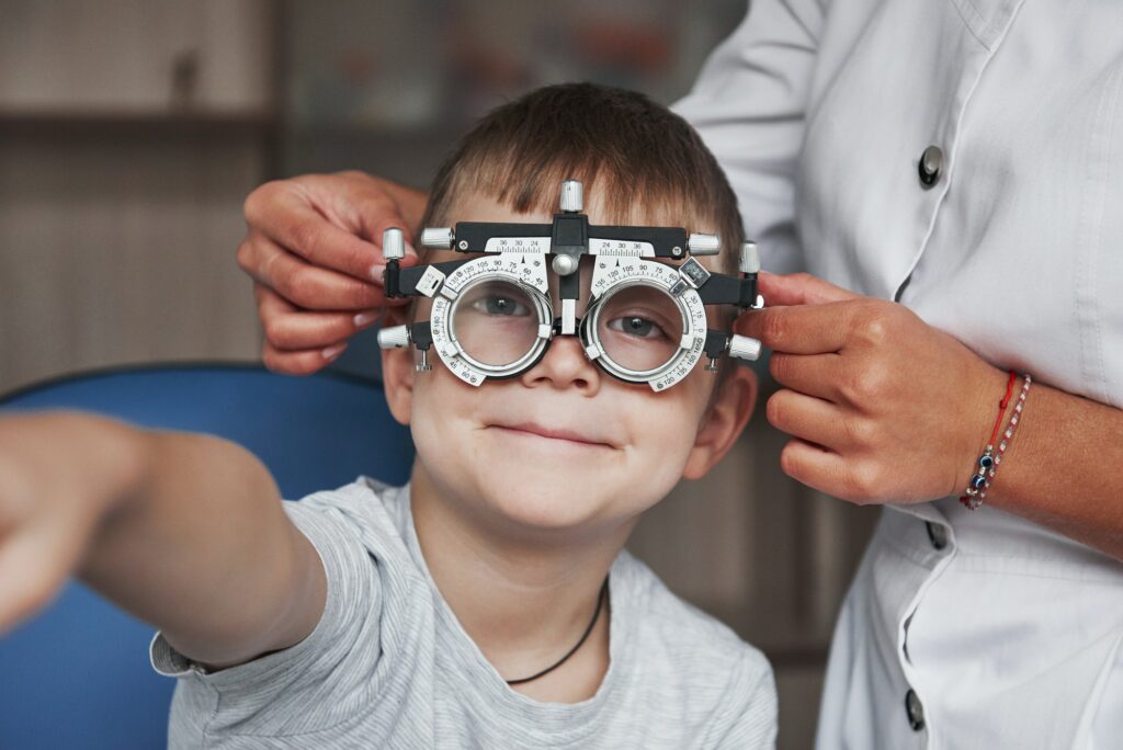 Kid smiling and raising hand while undergoing eye test with phoropter